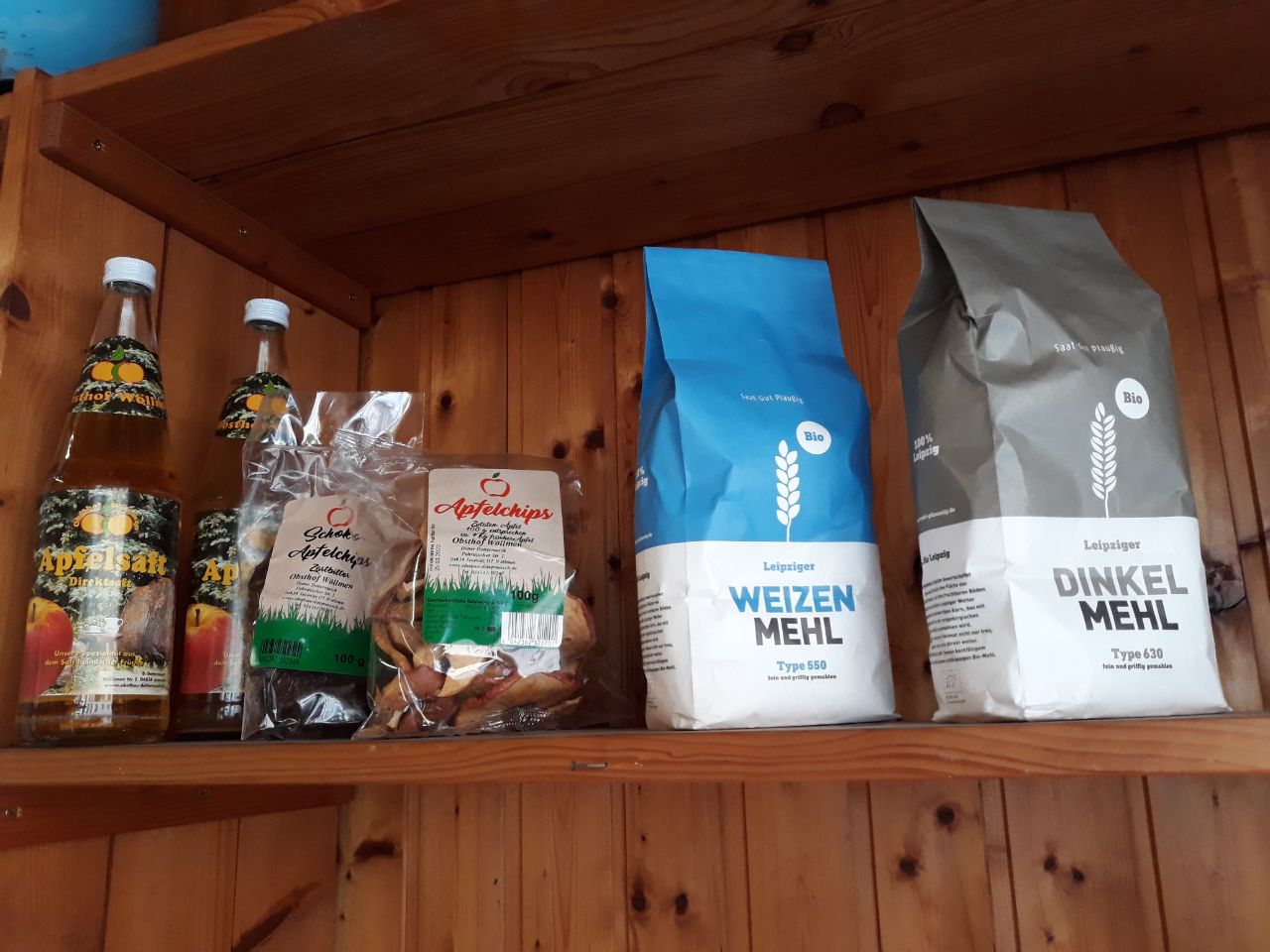 Das Bild zeigt die Anfänge des Regionalregals im Büro des Allmende Taucha e.V.. In einem Regalfach aus Holz stehen zwei Packungen Mehl, zwei Flaschen Apfelsaft und zwei Tüten Apfelchips. Die Produkte kommen Produzenten und Produzentinnen aus der Region um Leipzig.